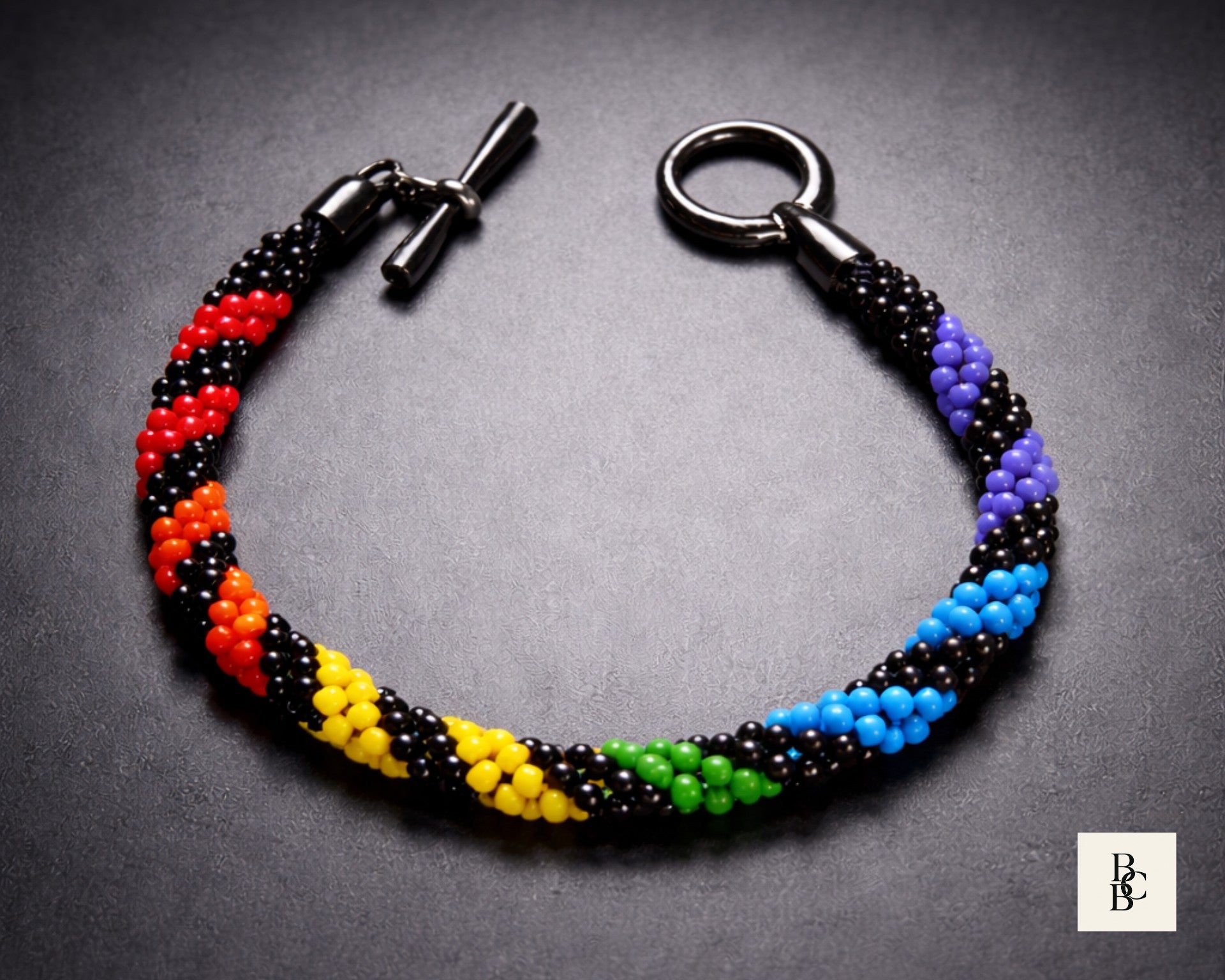 Rainbow Helix Pride Kumihimo bracelet with black glass seed beads, rainbow spiral accents, and a gunmetal-tone toggle clasp on a dark background.