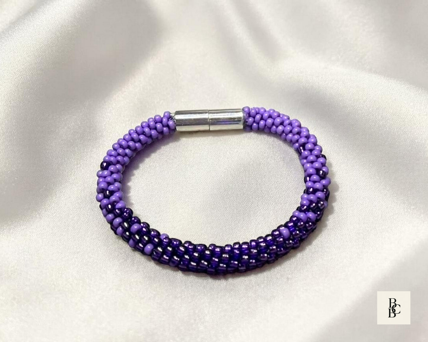 Shades of Violet Alzheimer’s Awareness Kumihimo bracelet displayed on ivory satin, featuring purple glass seed beads and a silver-tone magnetic clasp.
