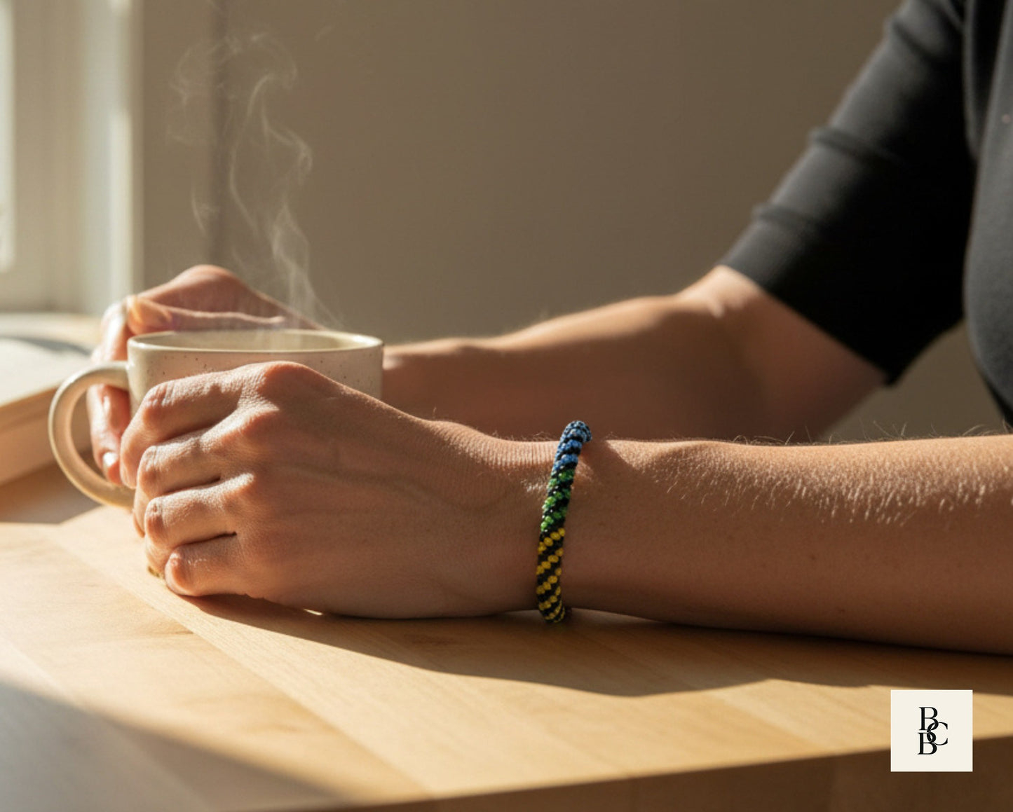 Rainbow Illusion beaded Pride bracelet worn while holding a warm mug, showcasing its black Kumihimo braid and bright rainbow pattern.