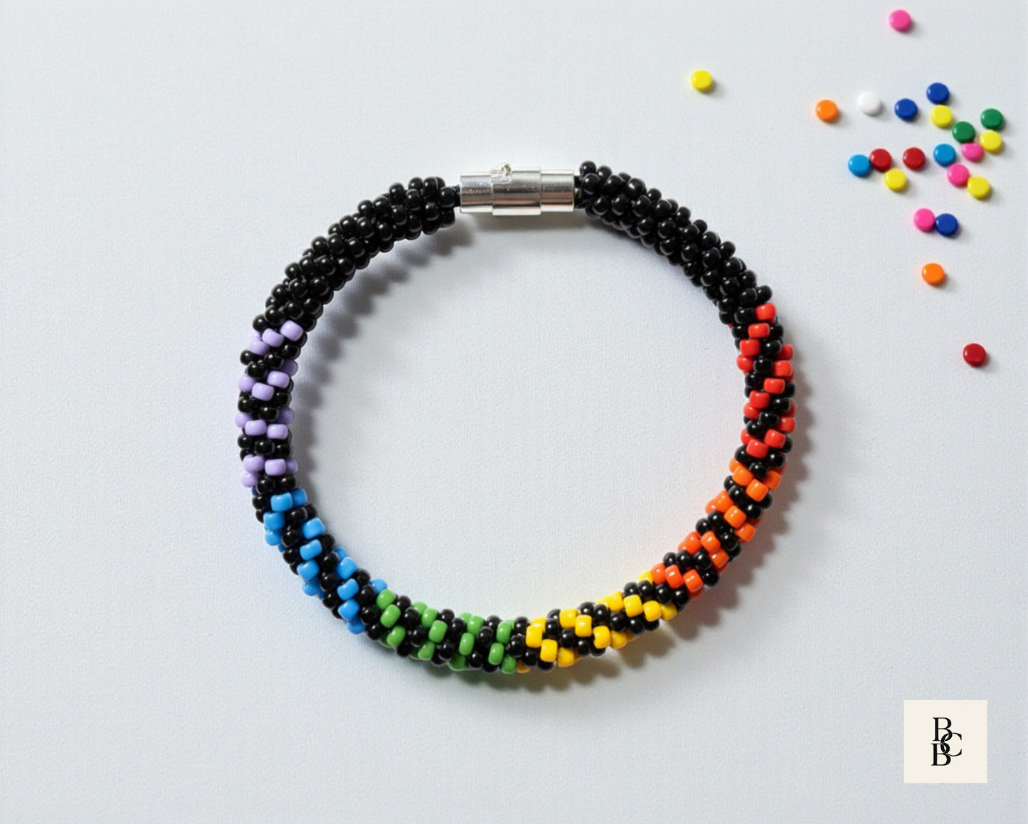 Flat lay of the Rainbow Illusion Pride Kumihimo bracelet with black and rainbow glass seed beads, silver clasp, and colorful bead accents.